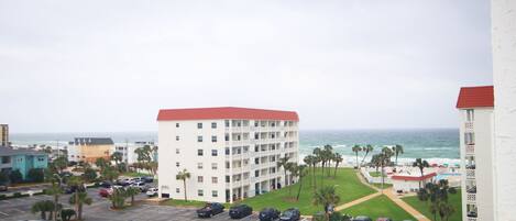 A multi-story hotel with a red roof, parking lot, and a view of the ocean.