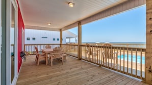 Eine Holzterrasse mit Tisch und Stühlen mit Blick auf ein Schwimmbecken und einen Strand.