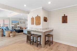 A modern kitchen with a white countertop, wooden stools, and a dining area with a sofa and coffee table. - Anster / 144 Tuna Drive 3 Bedroom Home by RedAwning (Supply)