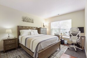 A bedroom with a bed, bedside table, lamp, desk, chair, and window with blinds. - River Pines C207 2 Bedroom Condo by RedAwning (Edwards)