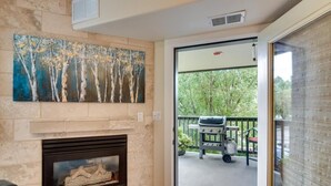 A fireplace with a stone mantle, a painting of trees, and a view of a patio with a grill and outdoor seating. - River Pines C207 2 Bedroom Condo by RedAwning (Edwards)