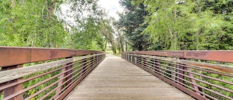 Un puente peatonal de madera con barandillas metálicas, rodeado de frondosa vegetación verde.