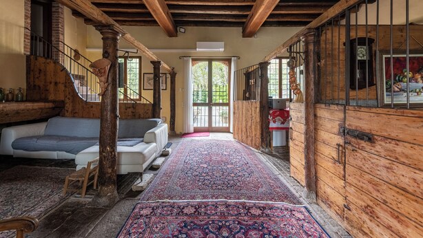 Un intérieur rustique avec des poutres apparentes, un canapé blanc, un tapis à motifs et une porte en bois ornée d’une horloge.