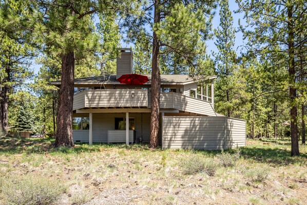 A two-story cabin with a covered porch and a red umbrella, surrounded by trees. - MCNA33 - Spacious Home with Great Views, AC, Hot Tub (Sunriver)