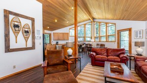 A spacious living area with a wooden ceiling, leather sofas, a wooden coffee table, and large windows offering views of the outdoors. - MCNA33 - Spacious Home with Great Views, AC, Hot Tub (Sunriver)