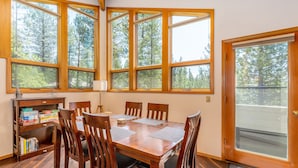 A dining area with a wooden table and chairs, large windows with a forest view, and a door leading outside. - MCNA33 - Spacious Home with Great Views, AC, Hot Tub (Sunriver)
