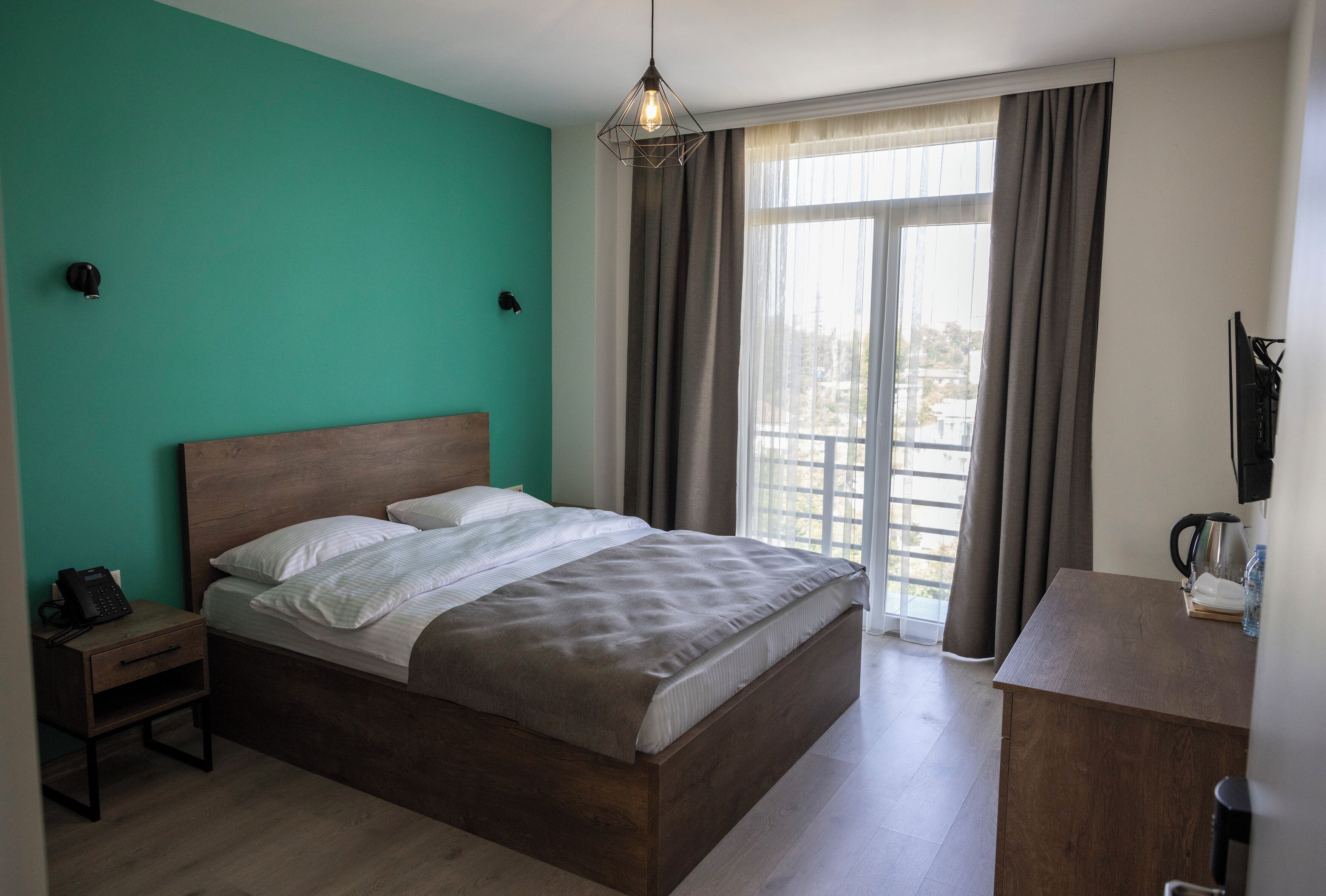 Un dormitorio con cama, mesita de noche, escritorio, silla, ventana con cortinas y vistas al exterior.