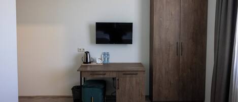 A room with a wooden desk, a television mounted on the wall, a wardrobe, and a window with curtains.