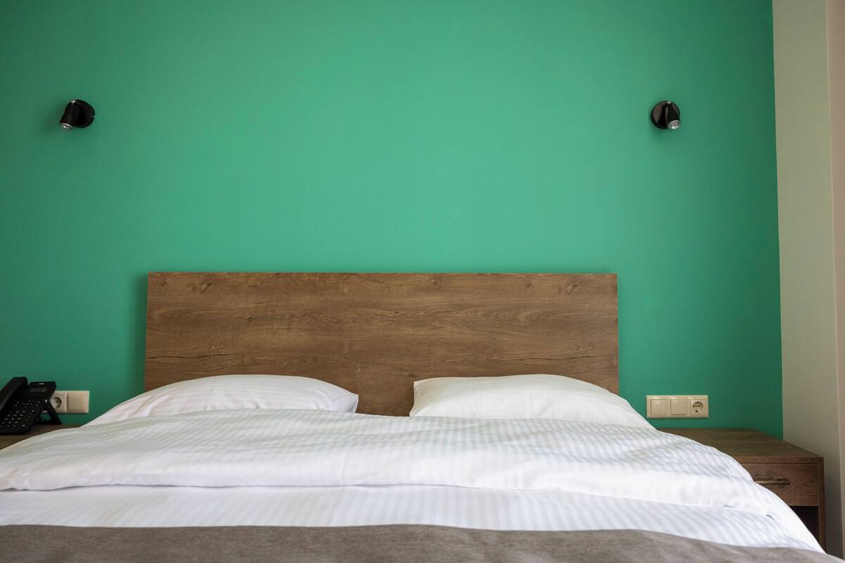 Una cama con ropa blanca, un cabecero de madera y dos lámparas de noche frente a una pared verde azulado.