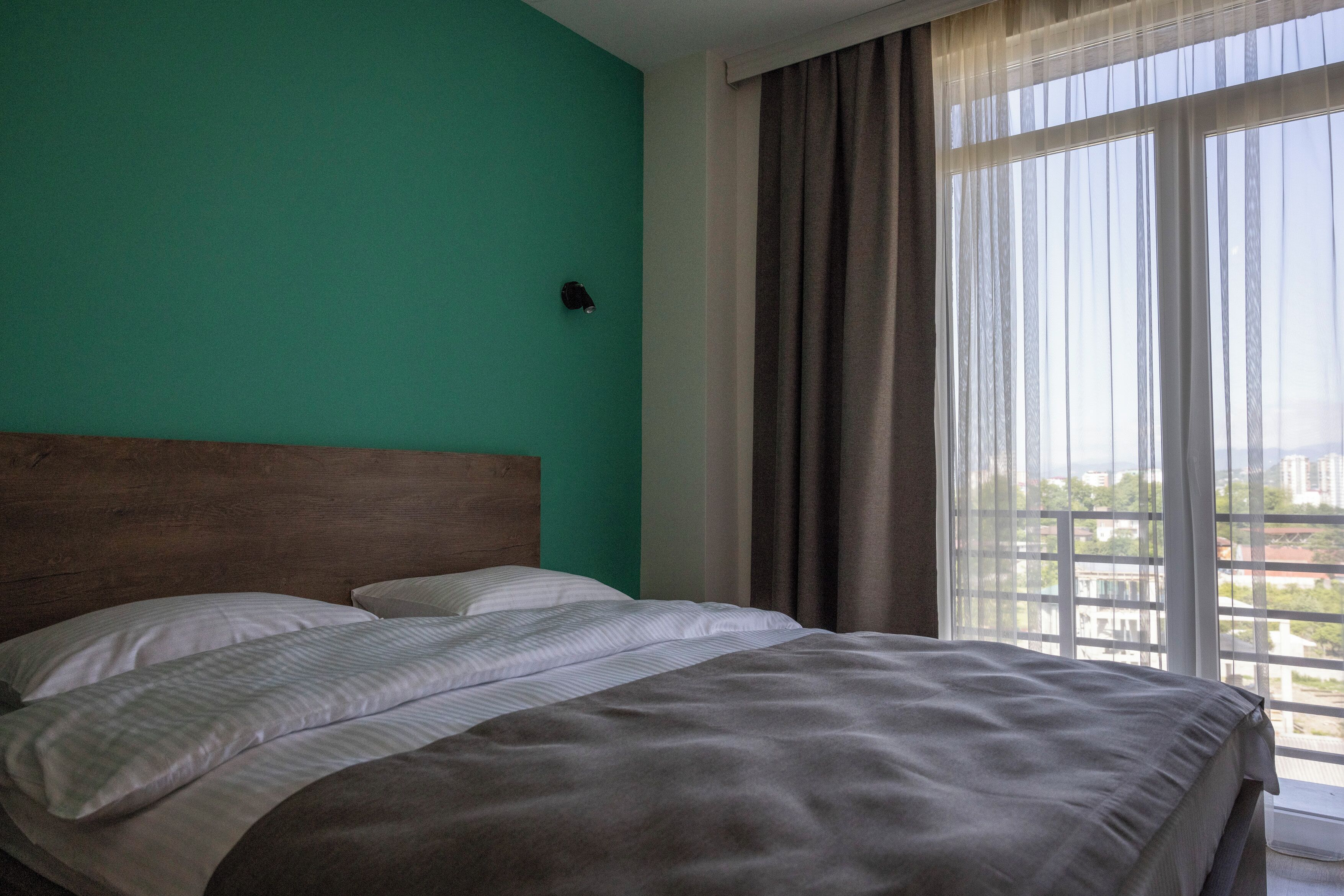 A hotel room with a bed, a headboard, a window with curtains, and a view of buildings outside.