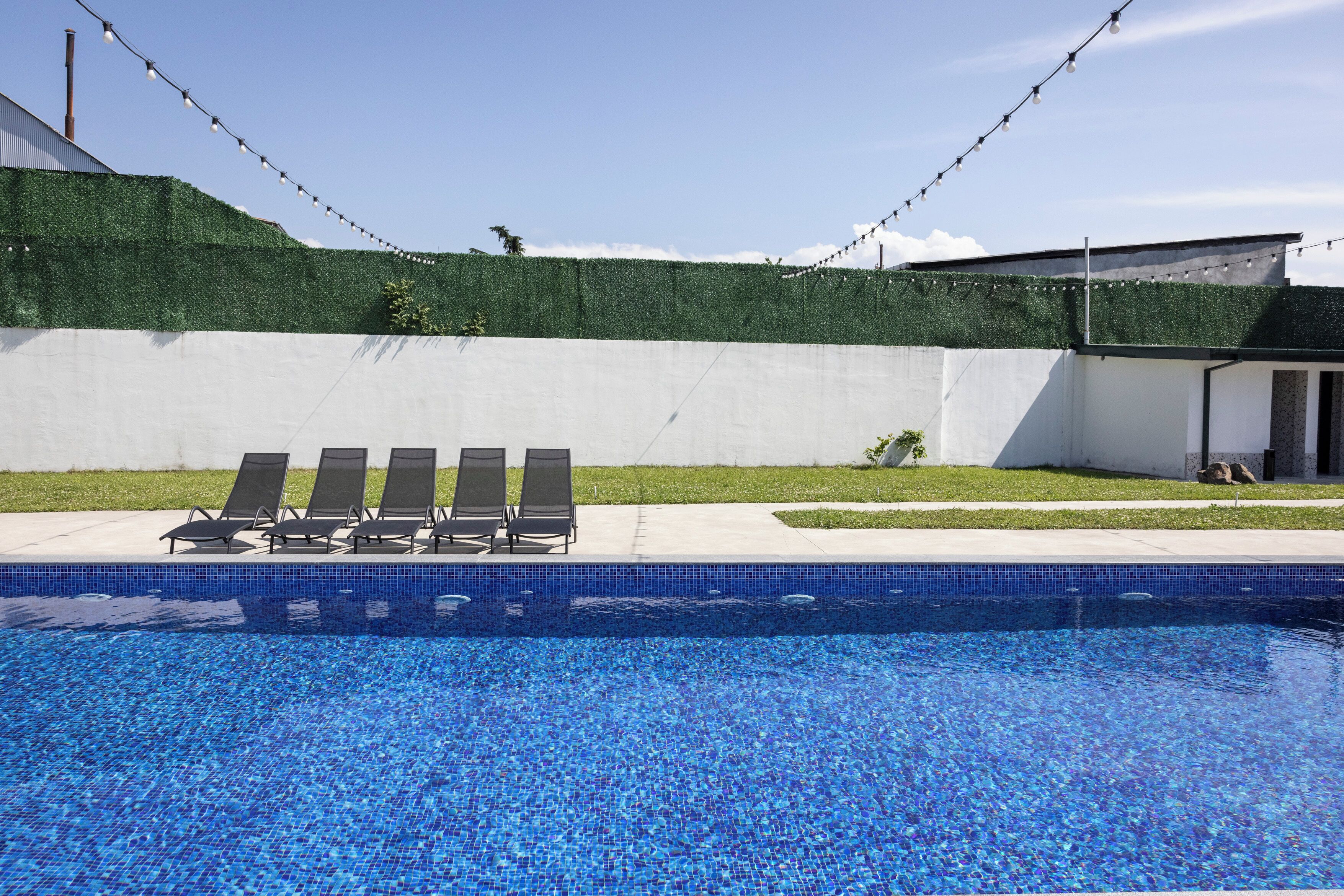 Ein Swimmingpool mit blauen Mosaikfliesen, umgeben von einer weißen Mauer und einem grünen Heckenstreifen.