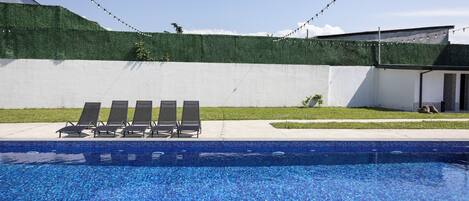 Ein Swimmingpool mit blauen Mosaikfliesen, umgeben von einer weißen Mauer und einem grünen Heckenstreifen.