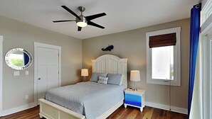 A bedroom with a bed, a ceiling fan, a bedside table with a lamp, a window with blinds, and a door. - Shore Sounds Fun | Beach Views + Pet Friendly! (Surf City)