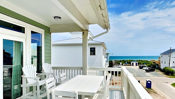 A covered outdoor area with a white table and chairs, a view of the ocean, and a parking area in the background. - Shore Sounds Fun | Beach Views + Pet Friendly! (Surf City)