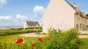 Een huis aan de kust met een witte gevel en een grijs dak, omgeven door een grindpad en weelderige begroeiing.