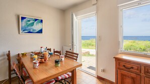 Een eetruimte met een houten tafel, stoelen en een schilderij aan de muur. Door een glazen deur is er uitzicht op de zee.