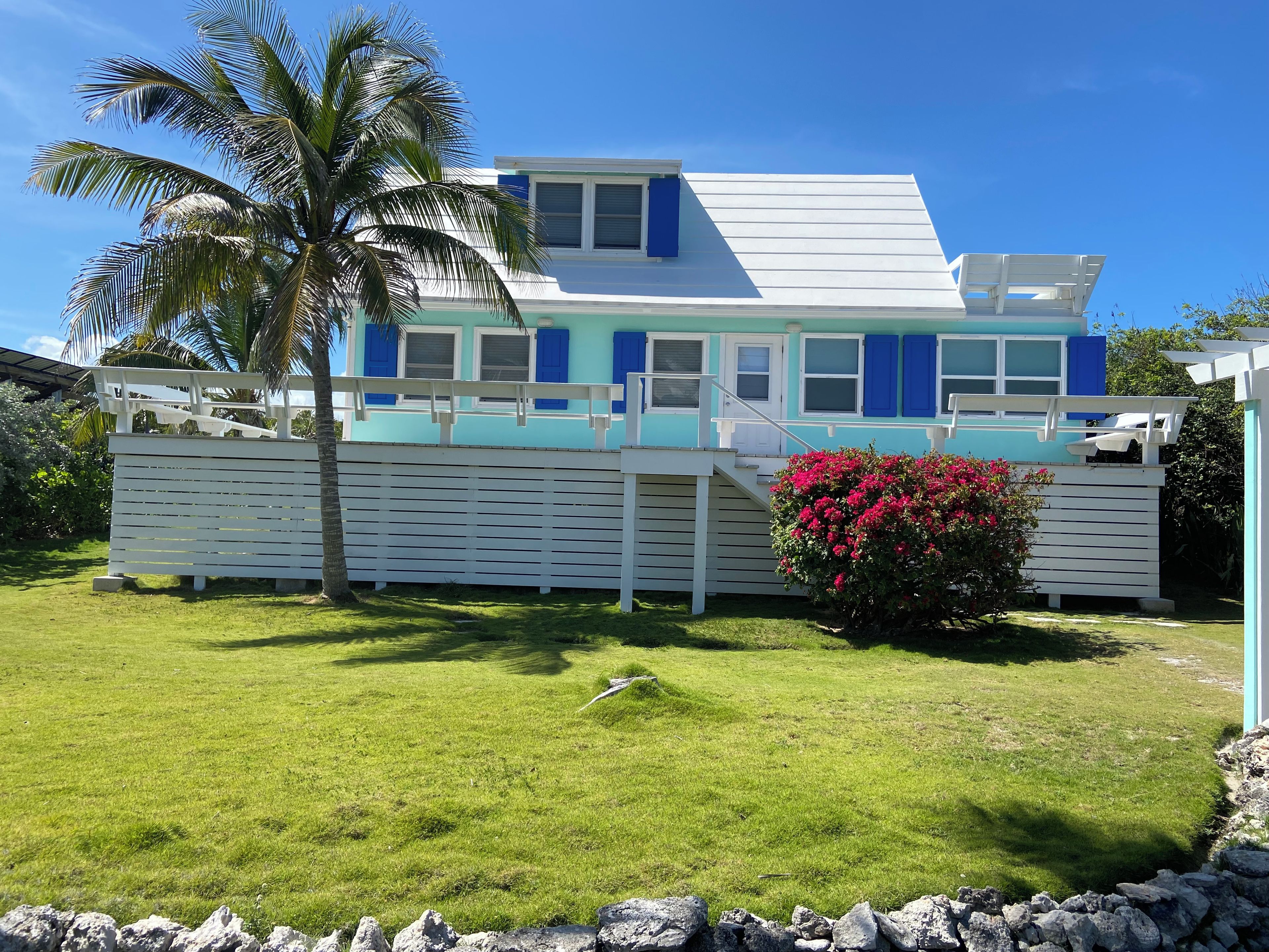 Une maison à deux étages, à l’extérieur blanc et bleu, avec un porche et un petit jardin agrémenté d’un palmier et de fleurs roses.