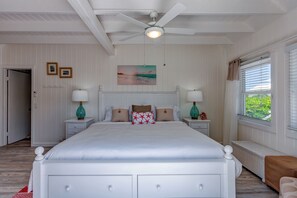Ein Schlafzimmer mit einem großen Bett, zwei Nachttischen mit Lampen, einem Deckenventilator, einem Fenster mit Jalousien und einem Wandbild.