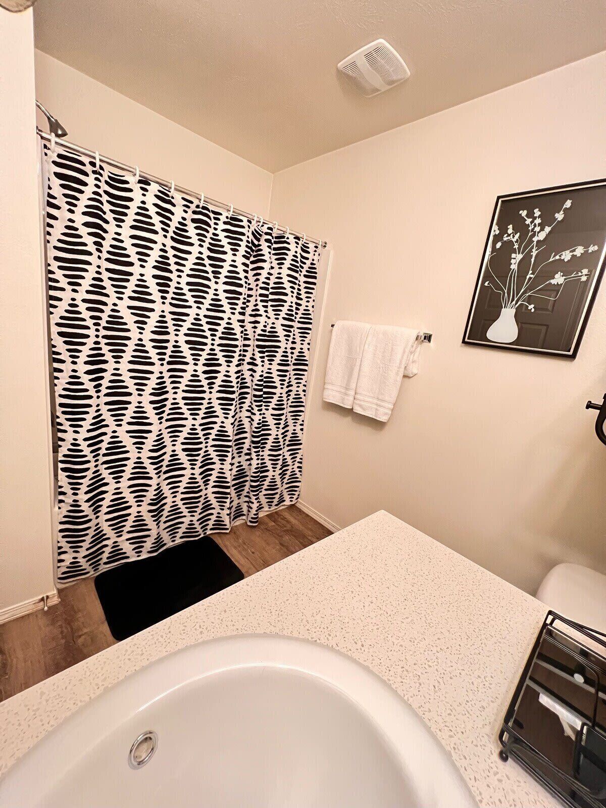 A bathroom with a bathtub, a shower curtain with a black and white pattern, a white towel on the wall, and a framed picture of a vase with flowers.