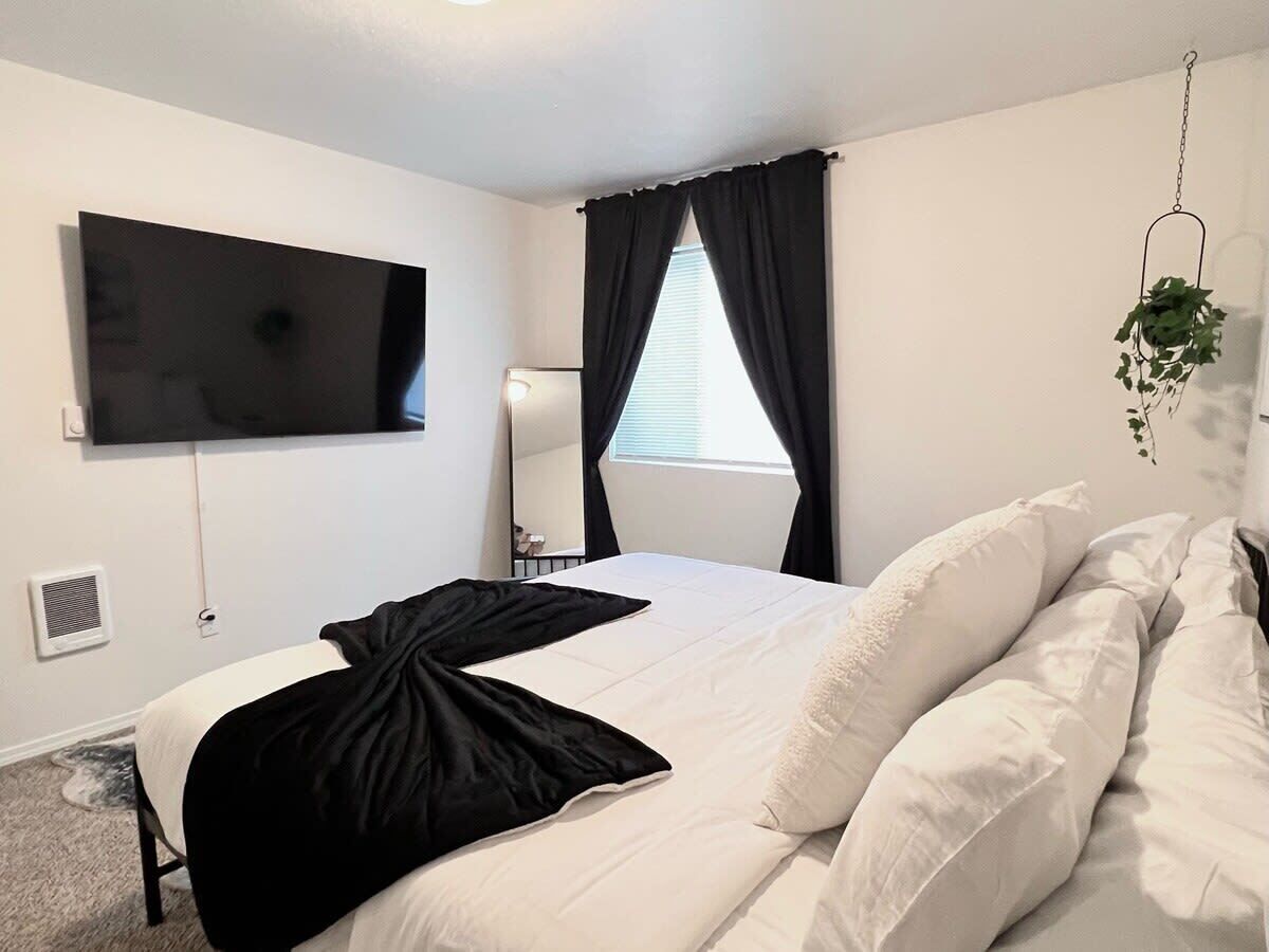A bedroom with a bed, a flat-screen TV mounted on the wall, a window with curtains, and a hanging plant.