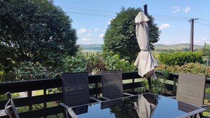 Ein Sitzbereich im Freien mit Tisch und Stühlen, umgeben von üppiger Vegetation und einem schönen Blick auf Hügel und See.