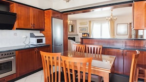 A modern kitchen with wooden cabinets, a built-in microwave, and a dining area with a glass table and wooden chairs. - Koridalos Terrace Apt w Spectacular View (Korydallos)