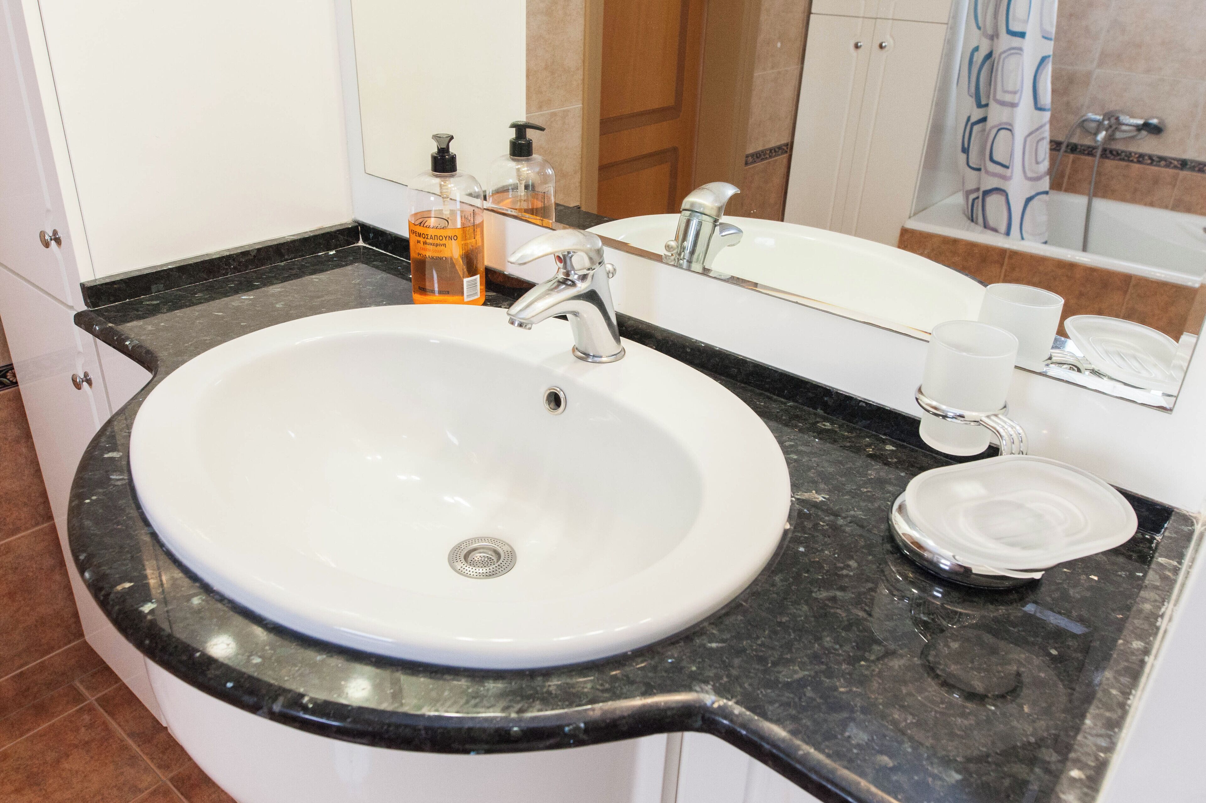 A bathroom with a white sink, black countertop, and a mirror above it. There are two bottles of soap and a cup on the countertop.