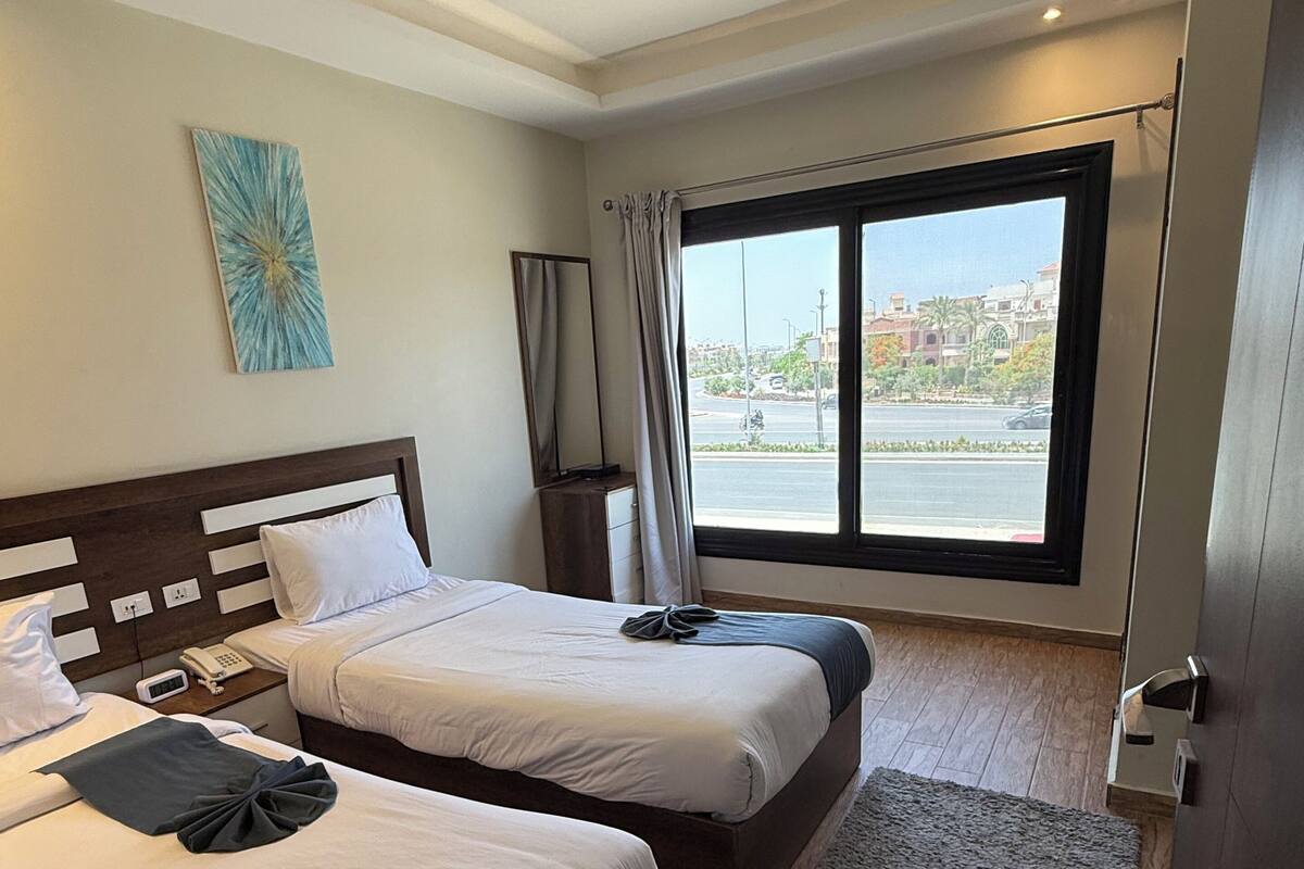 A hotel room with two beds, a large window, a grey rug, and a painting on the wall.