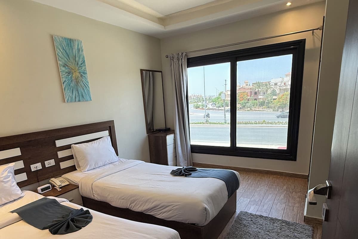 Habitación de hotel con dos camas, un ventanal grande, una alfombra gris y un cuadro en la pared.