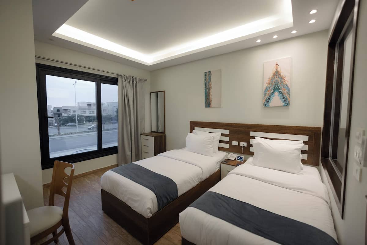A hotel room with two beds, a desk, a chair, a window with a view of the street, and artwork on the wall.