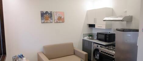 A compact living space with a sofa, a small coffee table with fruit, a microwave, a stove, and a washing machine.