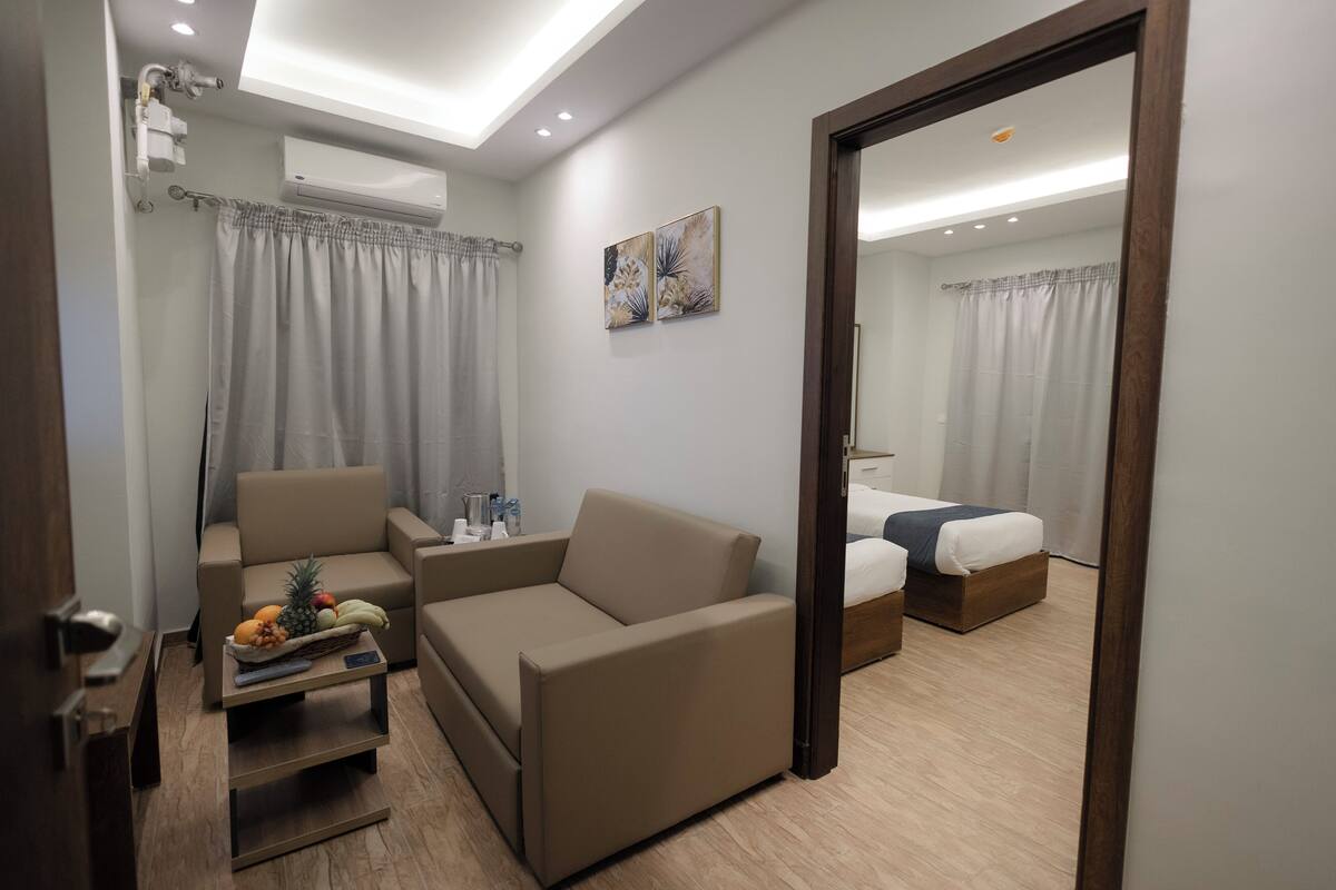 A modern hotel room with a sofa, a bed, a small table with fruits, and an air conditioning unit.