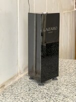 Un objeto rectangular metálico negro con el texto 'LAZARO SCENT', colocado sobre un suelo moteado frente a una pared lisa.