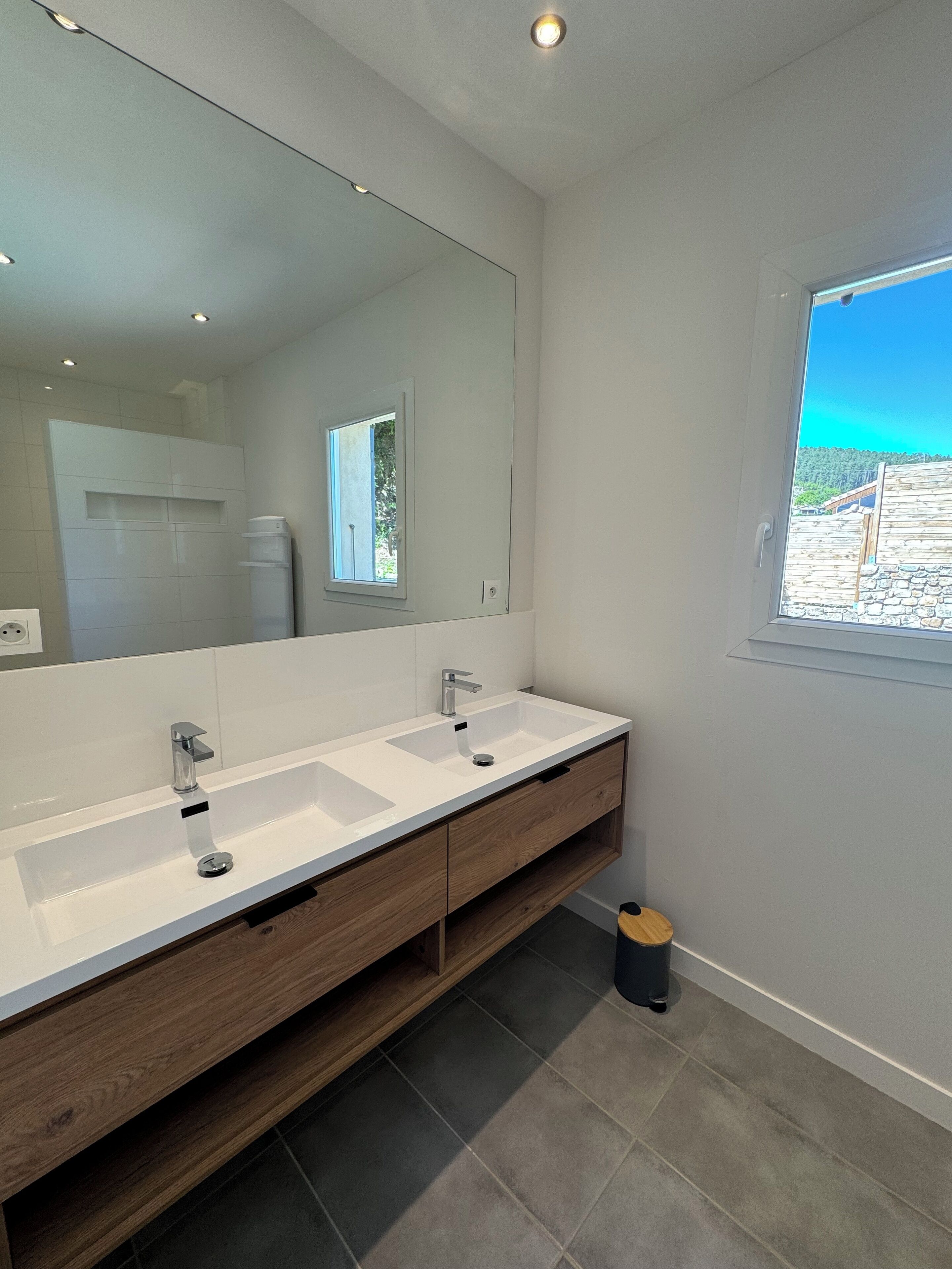 Une salle de bain moderne avec un grand miroir, deux vasques, un meuble-lavabo en bois et une fenêtre donnant sur un beau paysage.