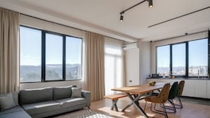 Een moderne woonkamer met een bank, eettafel en stoelen, grote ramen met gordijnen en uitzicht op de stad.