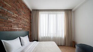 Een moderne slaapkamer met een bakstenen muur, een groot bed met witte lakens, een houten vloer en uitzicht op het groen door dunne gordijnen.