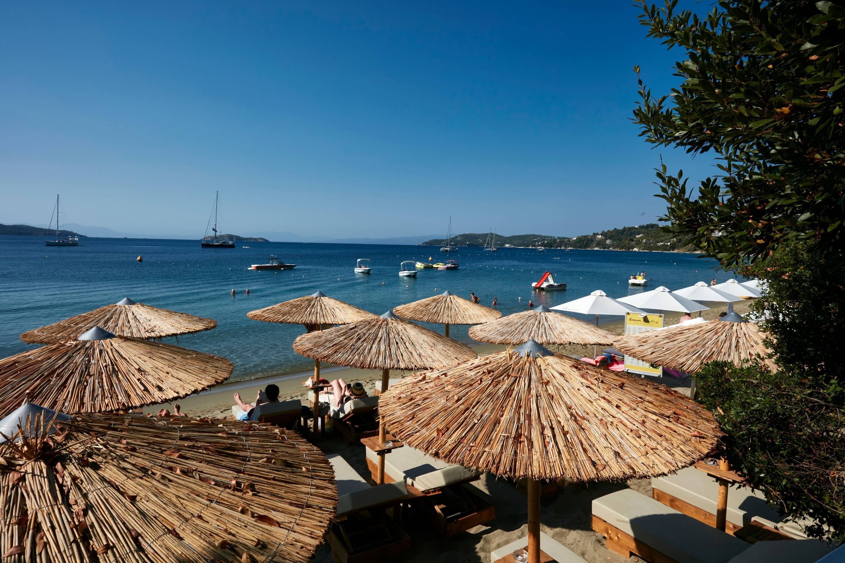Ein Strand mit Reetdachschirmen und Liegestühlen, Boote im Wasser und einem klaren Himmel.
