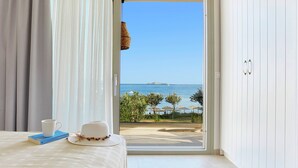 A room with a bed, a white hat, a cup, and a book on a table. There is a view of the sea and a beach through a glass door. - Skiathos Ammos Luxury Apartments in Megali Ammos Beach (Skiathos)
