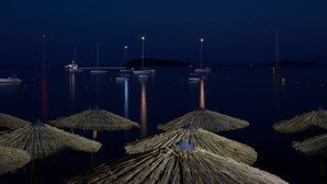 A nighttime marina with boats anchored in calm waters, illuminated by distant lights, and straw umbrellas in the foreground. - Skiathos Ammos Luxury Apartments (Skiathos)