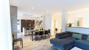 Un salon moderne avec un canapé d’angle, un coin repas avec une table et des chaises, et une cuisine en arrière-plan.