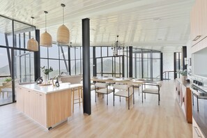 A spacious kitchen with a large island, modern pendant lights, and large windows offering a view of the outdoors. - Walaa Kulu – A Villa by 98 Acres (Ella)