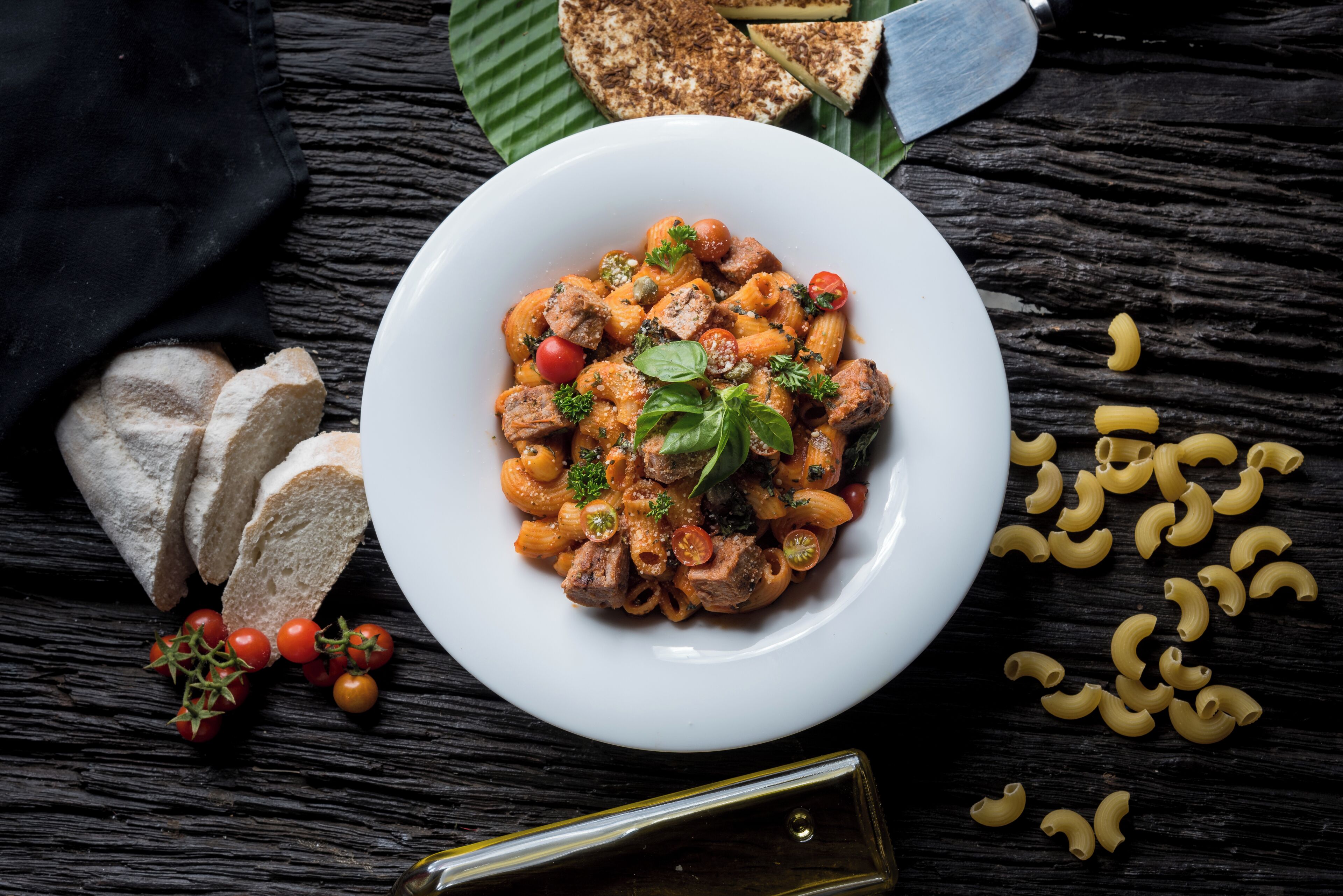 Un plato de pasta con carne y verduras, acompañado de pan y tomates sobre una mesa de madera oscura.