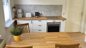 A compact kitchen with white cabinetry, a wooden countertop, a coffee maker, a kettle, and a toaster. There is a dining table with two chairs and a potted plant. - Ferienwohnung in Fehrenbach (Masserberg)