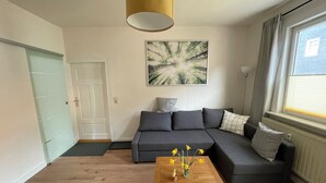 A modern living room with a sectional sofa, a wooden coffee table with flowers, a painting on the wall, and a window with curtains. - Ferienwohnung in Fehrenbach (Masserberg)