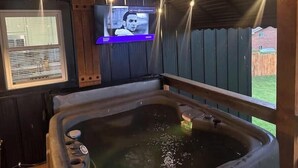 A room with a large bathtub, a television mounted on the wall, and a window with blinds.