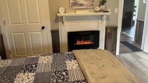A bedroom with a fireplace, a bed with a patterned bedspread, a painting above the mantel, and a door to the left.