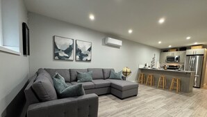 A modern living room with a grey sectional sofa, wooden flooring, and a kitchen area with a microwave, refrigerator, and stools.