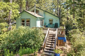 A small, light green cabin with a wooden deck and stairs leading to a porch. - Quiet Area, Perfect Couples Retreat Nestled in the Trees, Large Deck (Manzanita)