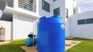 Ein blauer Wassertank und ein kleinerer, angeschlossener Tank mit einer Pumpe, gelegen auf einer Wiese mit einem weißen Gebäude im Hintergrund.