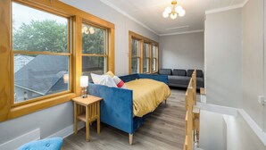 Ein Schlafzimmer mit einem blauen Schlafsofa, einem kleinen Tisch mit einer Lampe, einem grauen Sofa und großen Fenstern mit Holzrahmen.